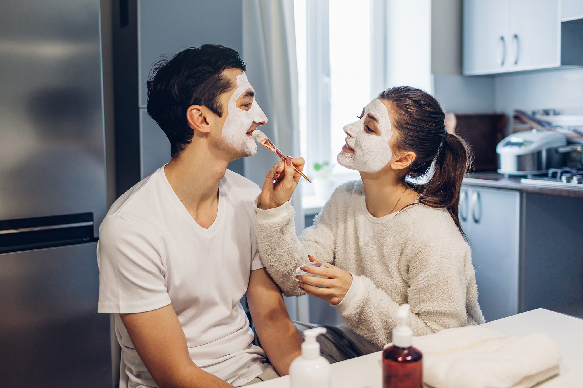 Woman applying clay mask on her boyfriends face. Young loving couple taking care of skin at home