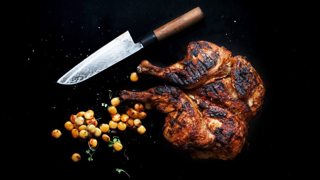 Poulet grillé en crapaudine sur fond noir. Couteau japonais et grains de maïs.