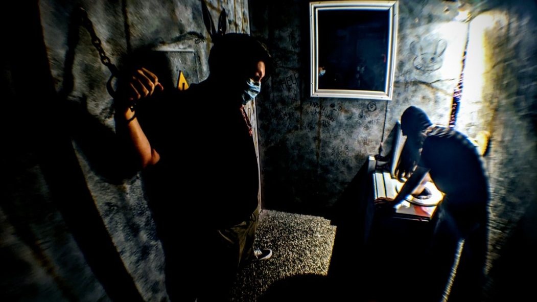 Un homme est enchaîné dans une pièce de jeu d'évasion.À sa droite un homme cherche dans une toilette. La pièce est sombre.