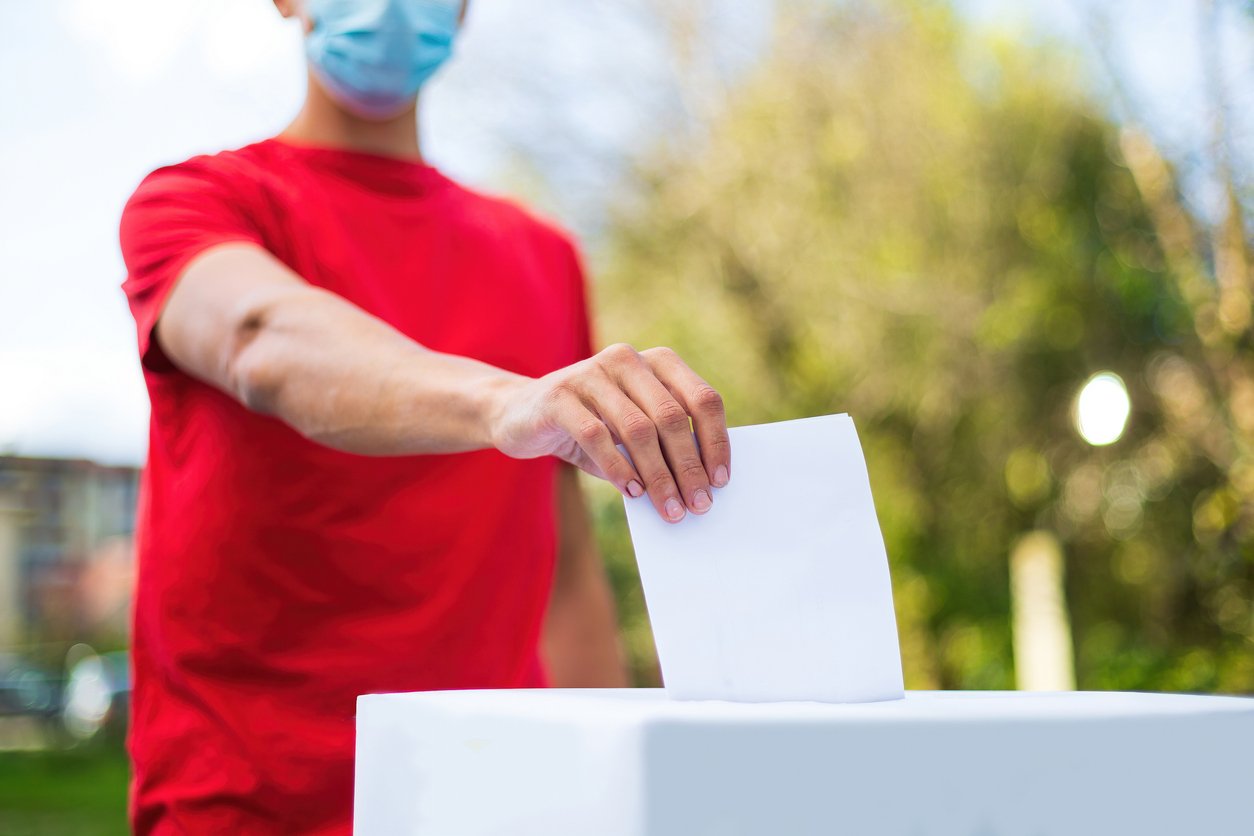Un homme en chandail rouge portant un masque chirurgical dépose son bulletin de vote dans une boîte de scrutin, à l'extérieur devant un paysage naturel.