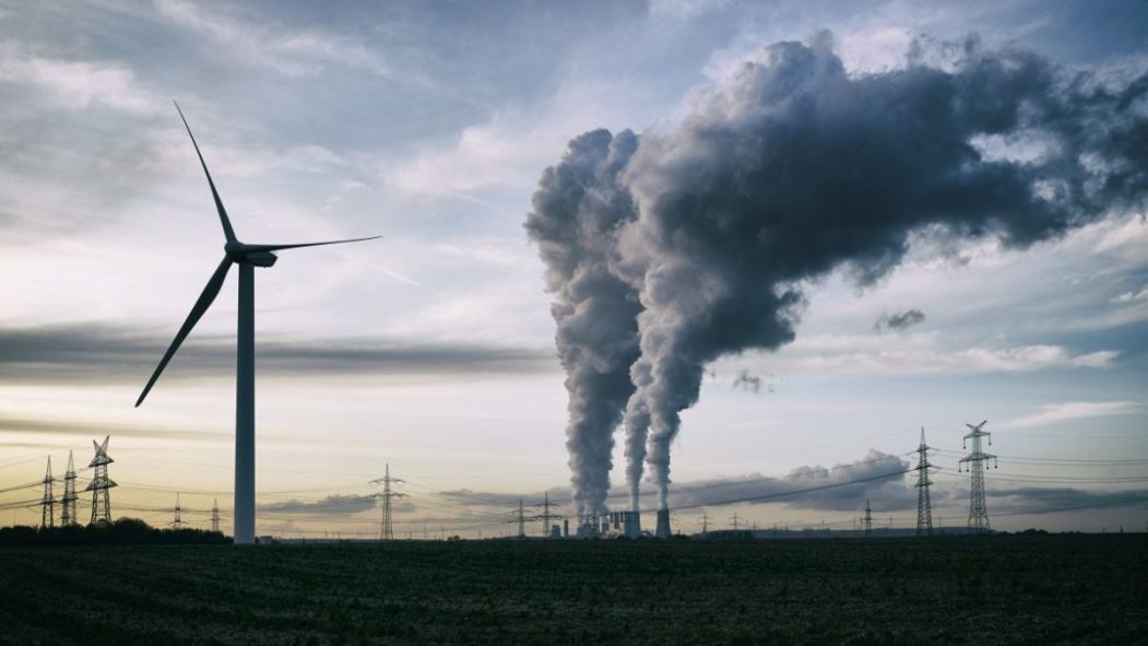 Une usine rejette de la fumée derrière un champ comprenant une éolienne.