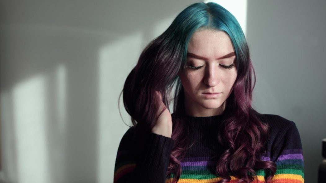 Une jeune femme avec un chandail à l'effigie de la communauté LGBTQ et des cheveux mauves et roses le regard baissé.