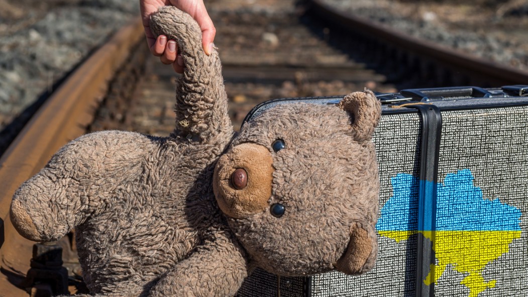 Une main représentant un enfant réfugié ukrainien tient un ours en peluche devant une valise montrant les couleurs et les frontières de l'Ukraine.