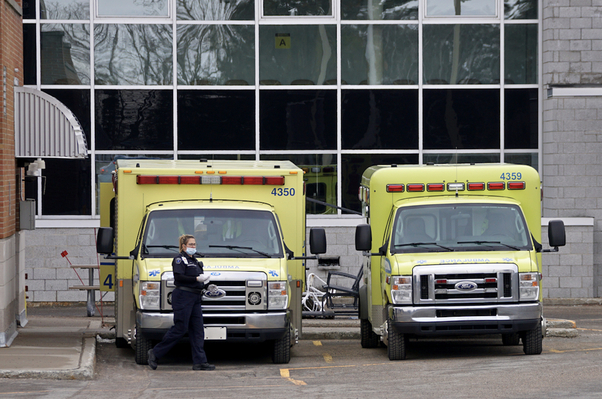 Deux ambulances jaunes stationnées à l’entrée d’urgence de l’hôpital avec une dame paramédicale portant un masque.