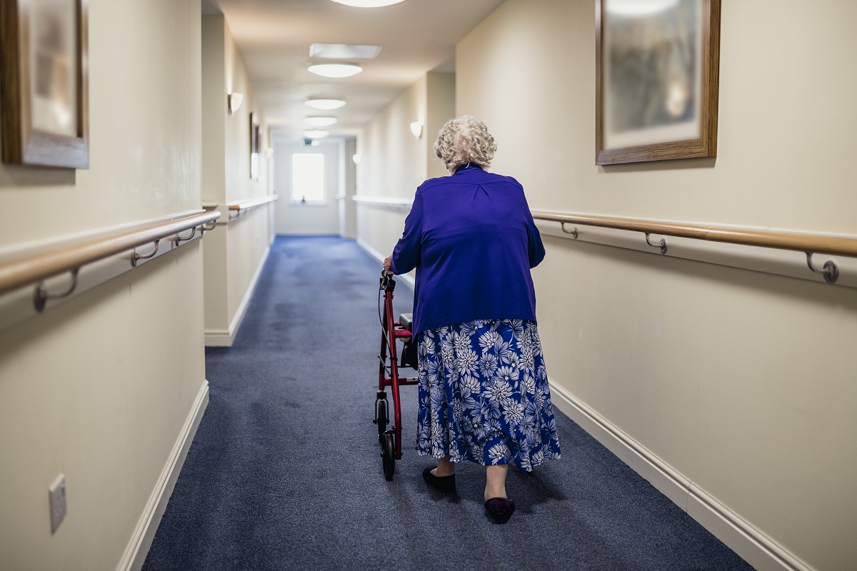 Une femme avec une marchette, de dos, dans un couloir