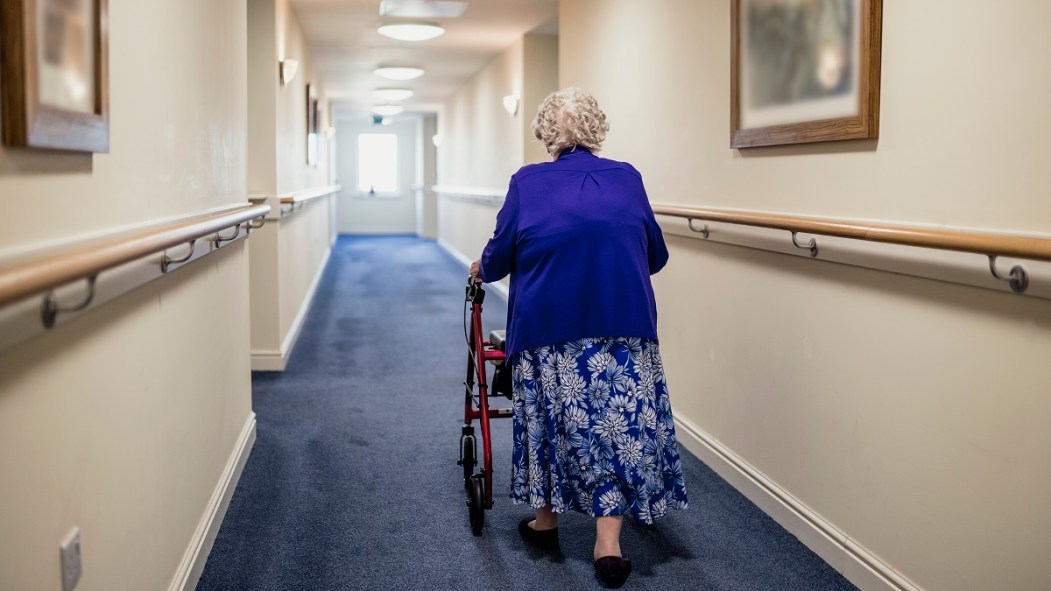 Une femme avec une marchette, de dos, dans un couloir